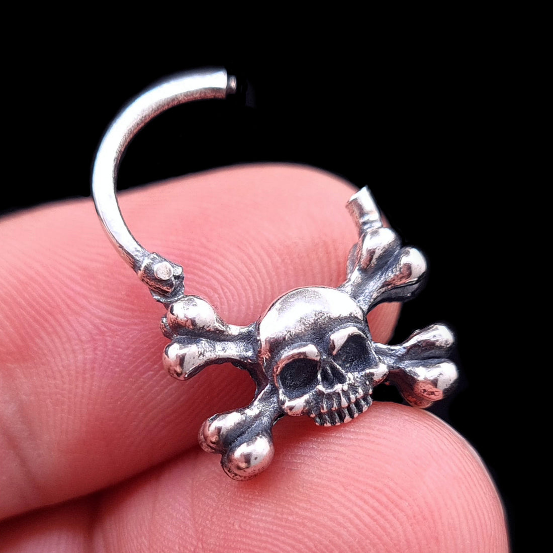 Sterling silver skull and crossbones septum clicker by RBZ Jewelry, shown with the hinge mechanism open and held by a hand to illustrate scale and the detailed, oxidized gothic design.
