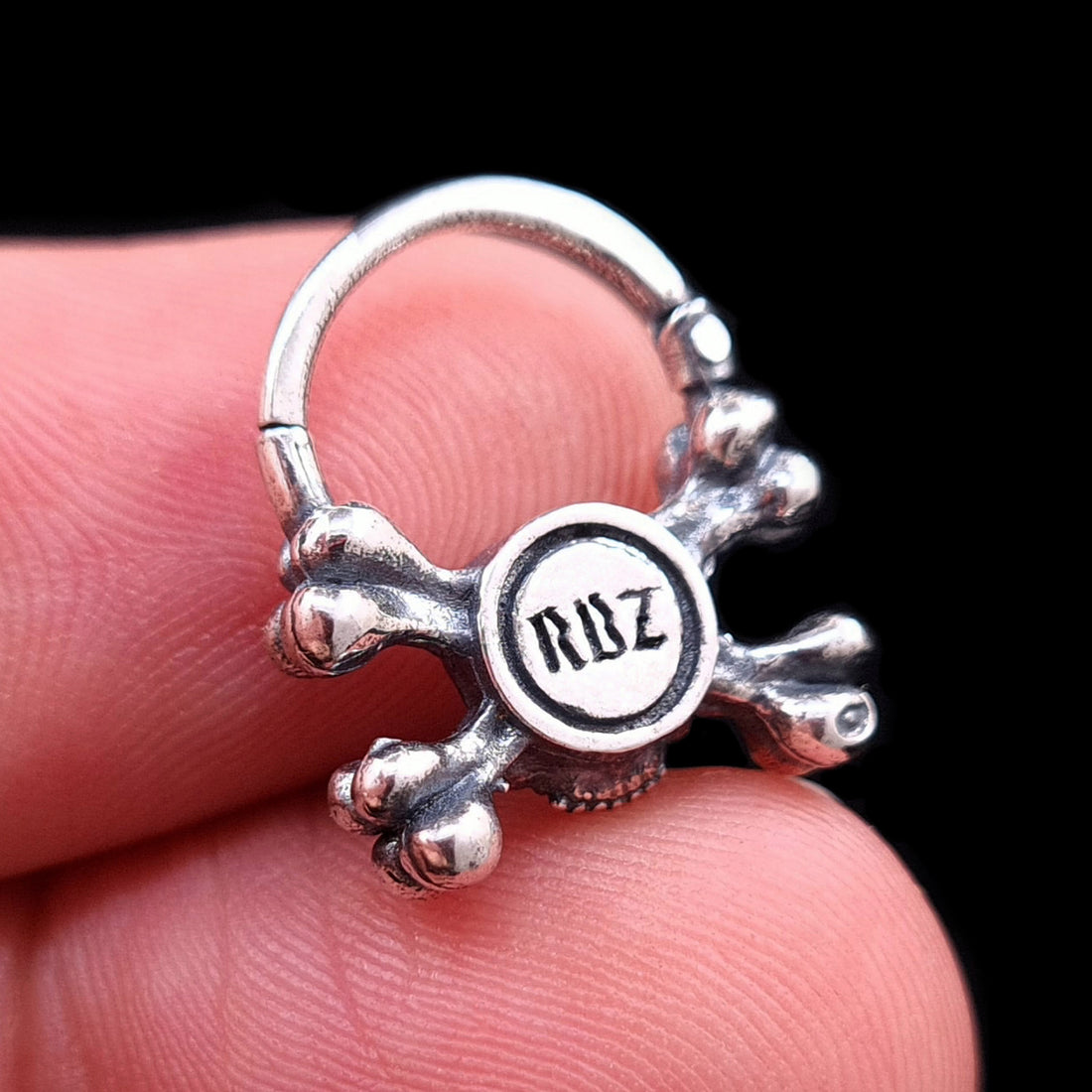 Back view of the oxidized sterling silver crossbones skull septum ring by RBZ Jewelry, held in a hand to demonstrate scale and featuring the gothic RBZ maker's mark engraved on the reverse.