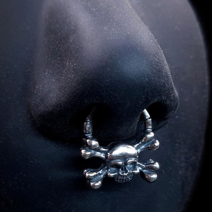 Sterling silver crossbones skull septum ring by RBZ Jewelry, displayed worn on a life-size black silicone nose to demonstrate fit and the bold, oxidized gothic memento mori design.