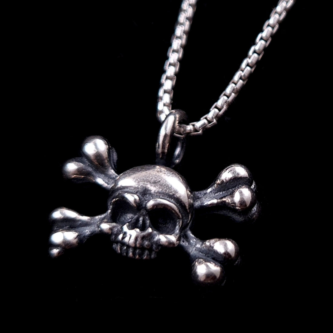 Handcrafted Gothic sterling silver skull and crossbones pendant by RBZ Jewelry, featuring intricate oxidized detailing and a polished finish, hanging from a silver box chain against a black background.