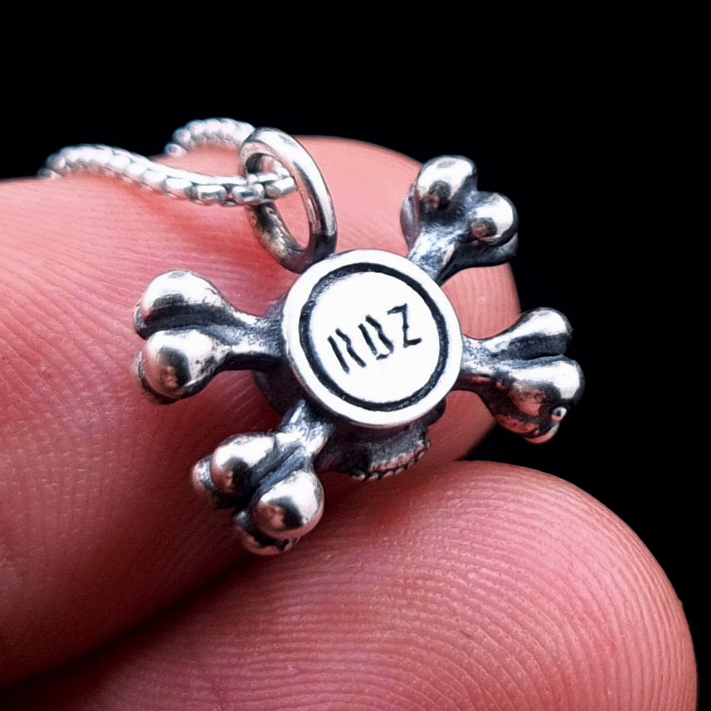 Back view of the sterling silver skull and crossbones pendant by RBZ Jewelry, showcasing the RBZ logo hallmark on a polished circular base, held between fingers to demonstrate its subtle, dainty scale.