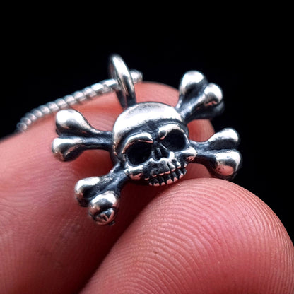 Detailed close-up front view of the RBZ Jewelry sterling silver Gothic skull and crossbones pendant being held between two fingers to show its scale. The image highlights the intricate oxidized detailing and polished finish, against a black background with a partial silver chain.