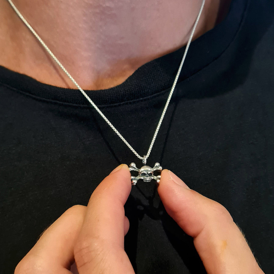 Lifestyle view of a male model wearing the RBZ Jewelry sterling silver skull and crossbones pendant, holding the piece between his fingers to showcase its dainty scale and intricate Gothic detailing against a black t-shirt.