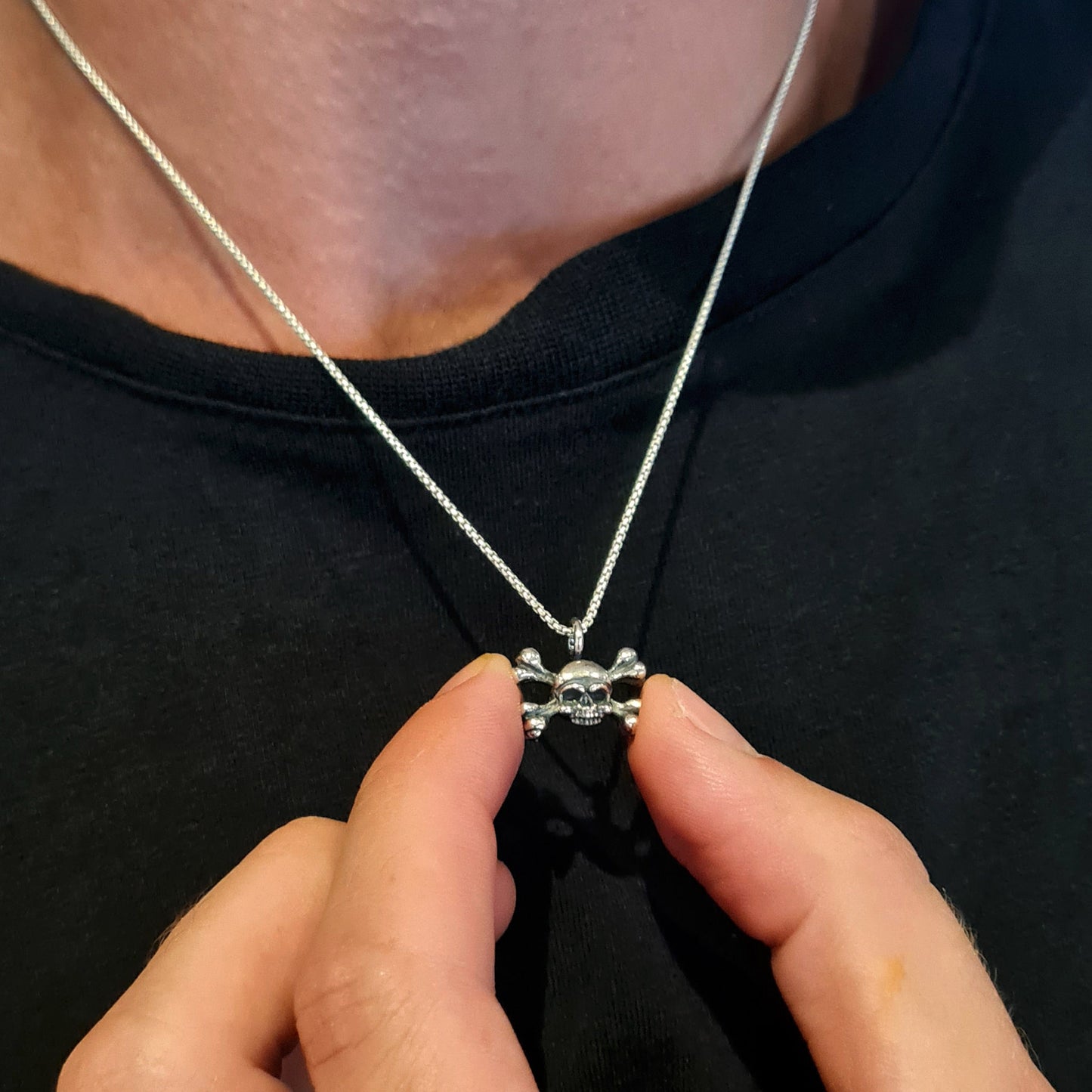 Lifestyle view of a male model wearing the RBZ Jewelry sterling silver skull and crossbones pendant, holding the piece between his fingers to showcase its dainty scale and intricate Gothic detailing against a black t-shirt.