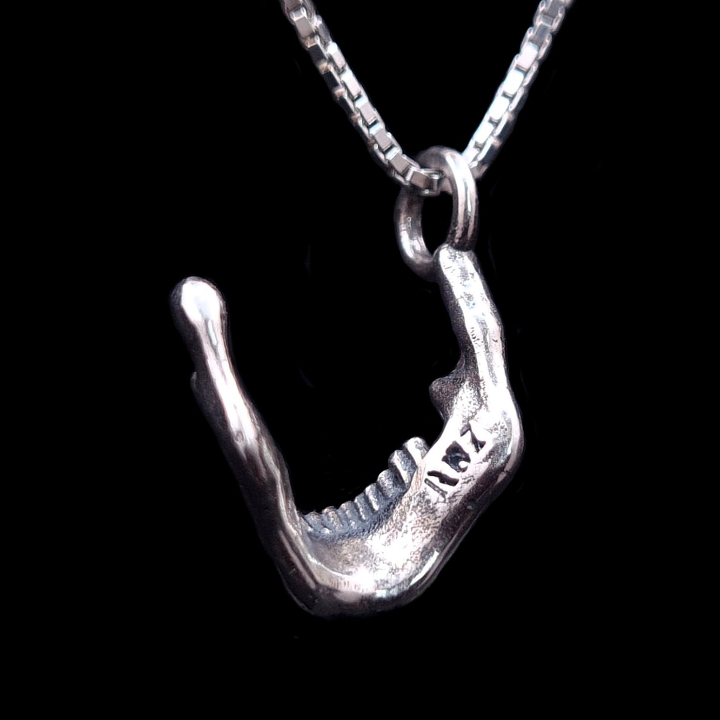 Rear view of the RBZ Jewelry oxidized sterling silver skull jaw pendant, highlighting the engraved RBZ logo on the inner mandible. The detailed gothic skeletal texture and Memento Mori design are shown against a black background, suspended from a silver chain.