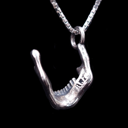 Rear view of the RBZ Jewelry oxidized sterling silver skull jaw pendant, highlighting the engraved RBZ logo on the inner mandible. The detailed gothic skeletal texture and Memento Mori design are shown against a black background, suspended from a silver chain.