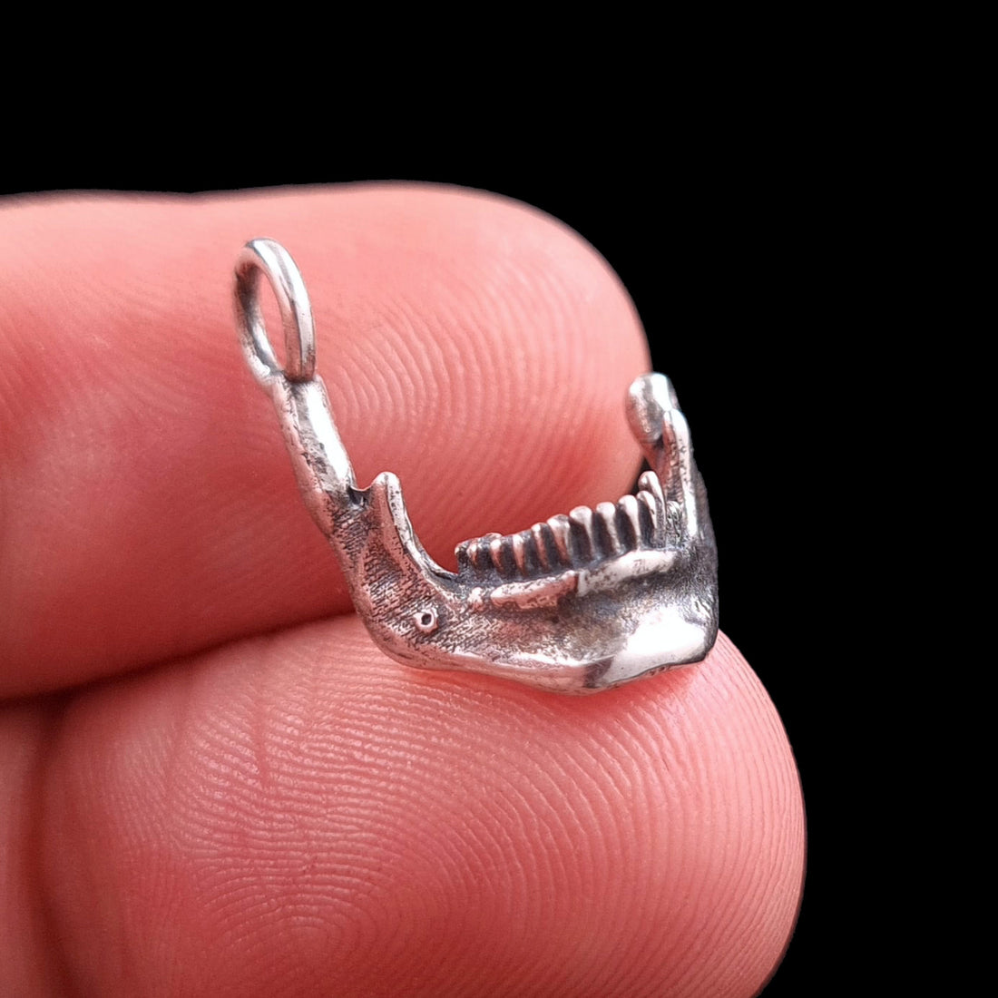 Handcrafted sterling silver skull jaw pendant by RBZ Jewelry held on a fingertip to provide a clear scale and size reference. The detailed gothic mandible, featuring an oxidized skeletal texture and Memento Mori design, is shown in profile facing right against a black background.