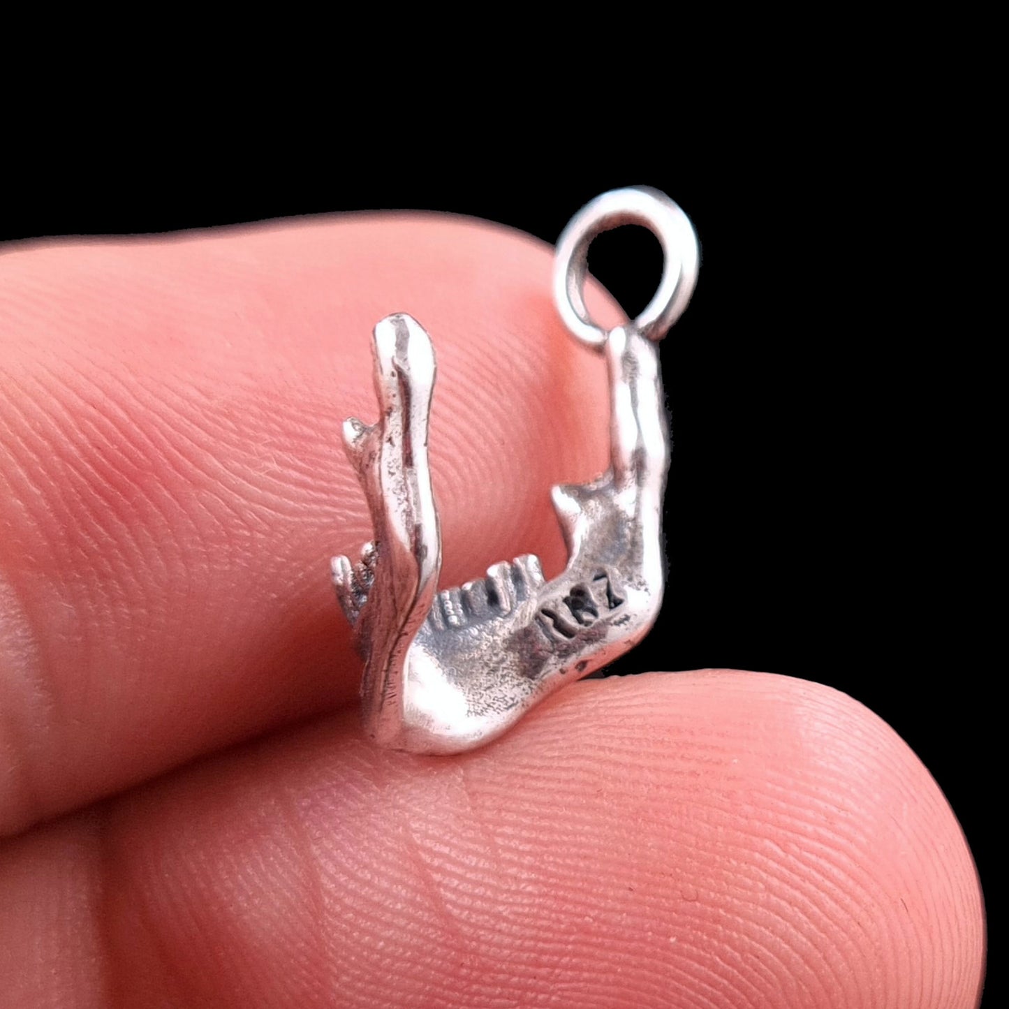 Handcrafted sterling silver skull jaw pendant by RBZ Jewelry held on a fingertip for scale, showing the rear view with the engraved RBZ logo on the inner mandible. The detailed gothic skeletal texture and oxidized finish of the Memento Mori charm are clearly visible against a black background.