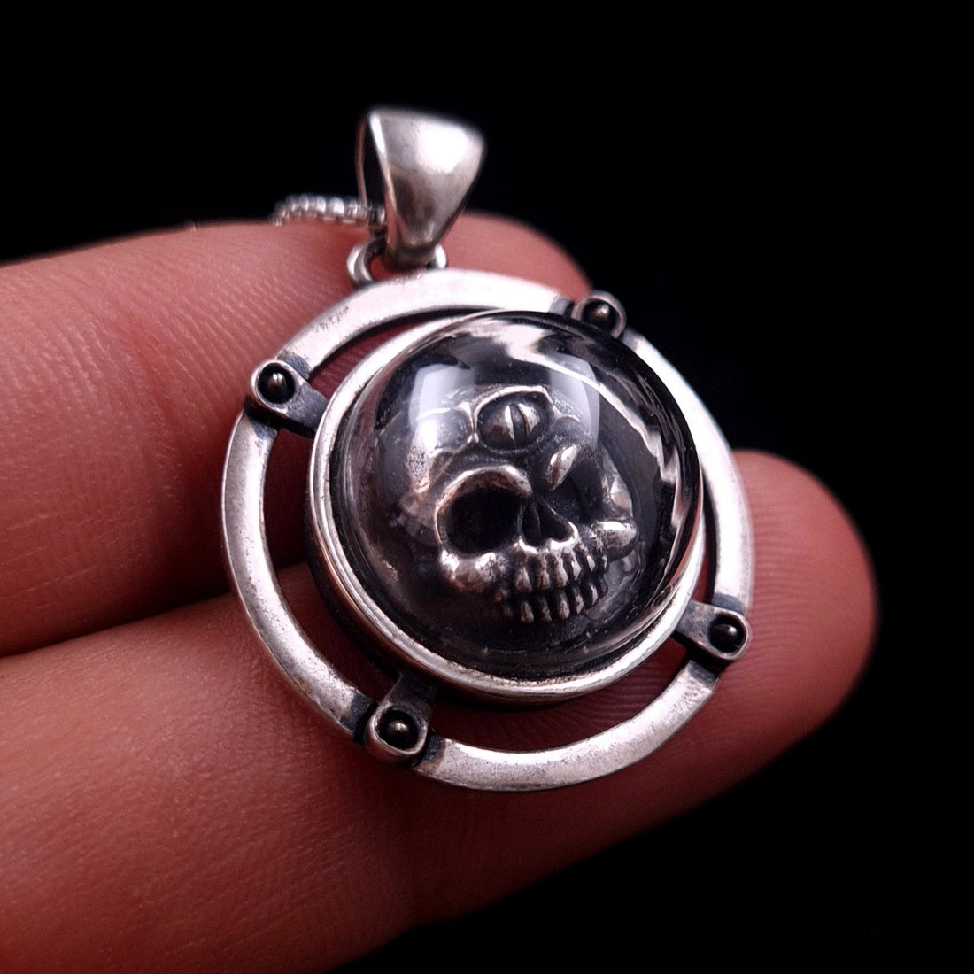 Scale view of the RBZ Jewelry sterling silver Third-eye Skull pendant held between two fingers, highlighting the intricate three-eyed anatomical skull protected within its clear glass dome and riveted silver housing.