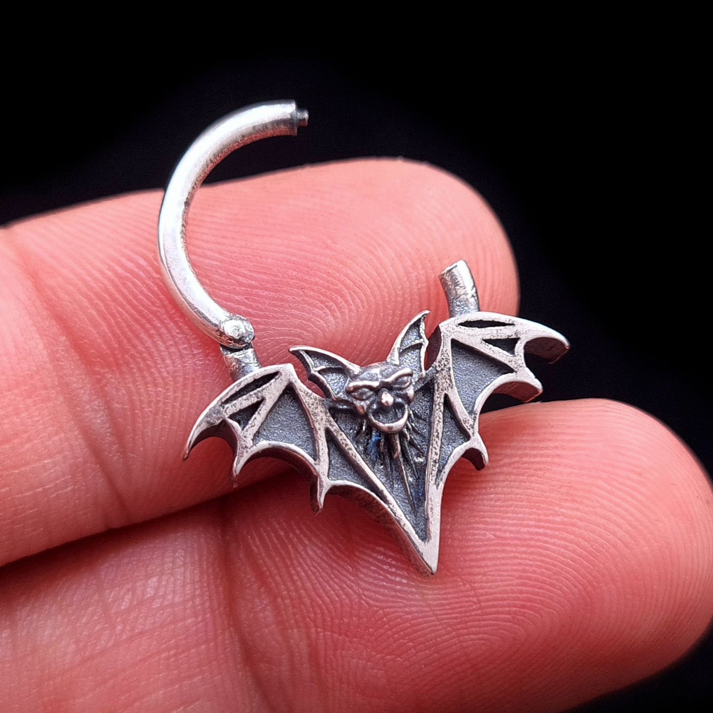 Handheld view of the sterling silver Gothic vampire bat septum ring by RBZ Jewelry in the open position, demonstrating the clicker hinge mechanism and scale against fingers.