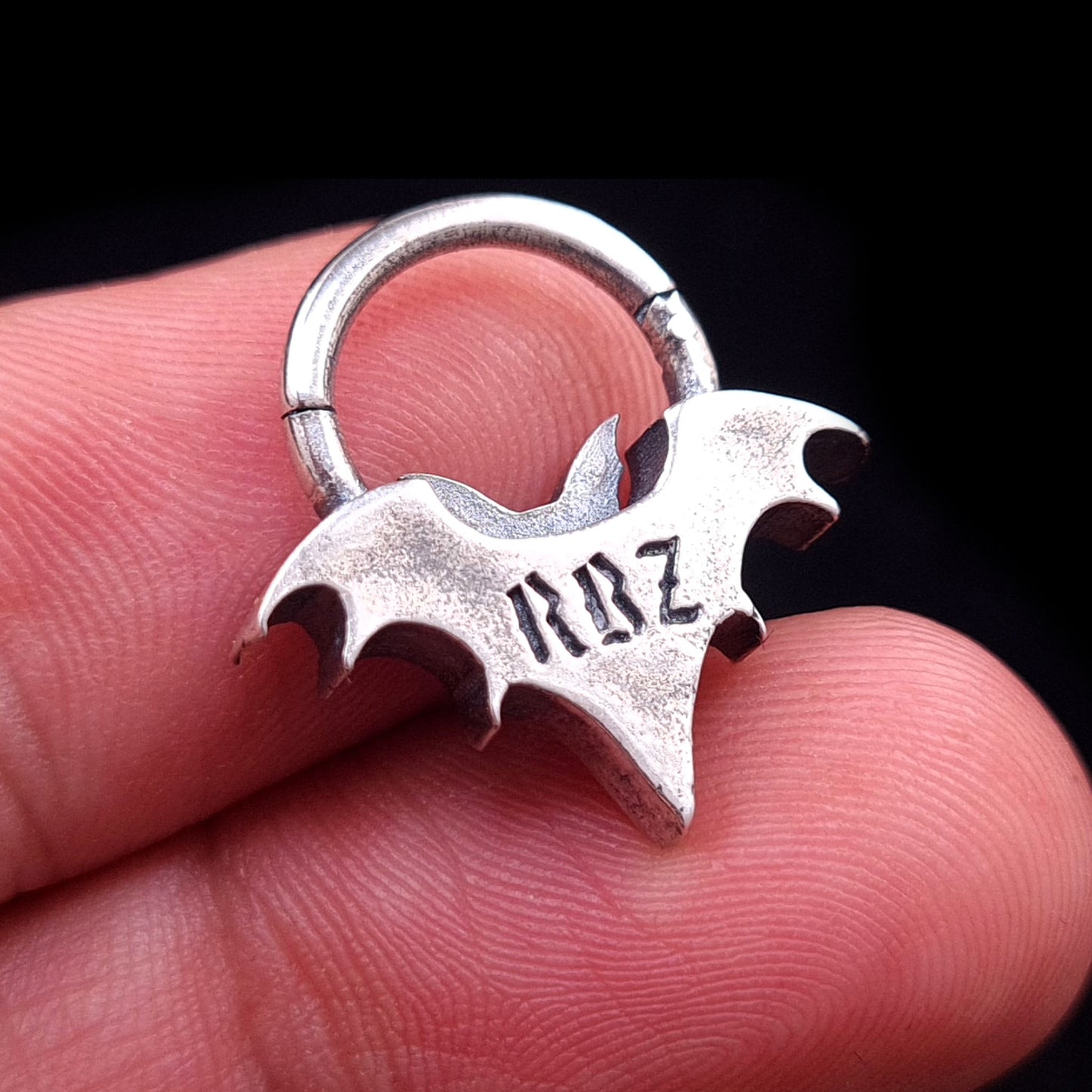 Back view of the sterling silver Gothic vampire bat septum clicker held in hand, showcasing the RBZ logo engraving and providing a sense of scale.