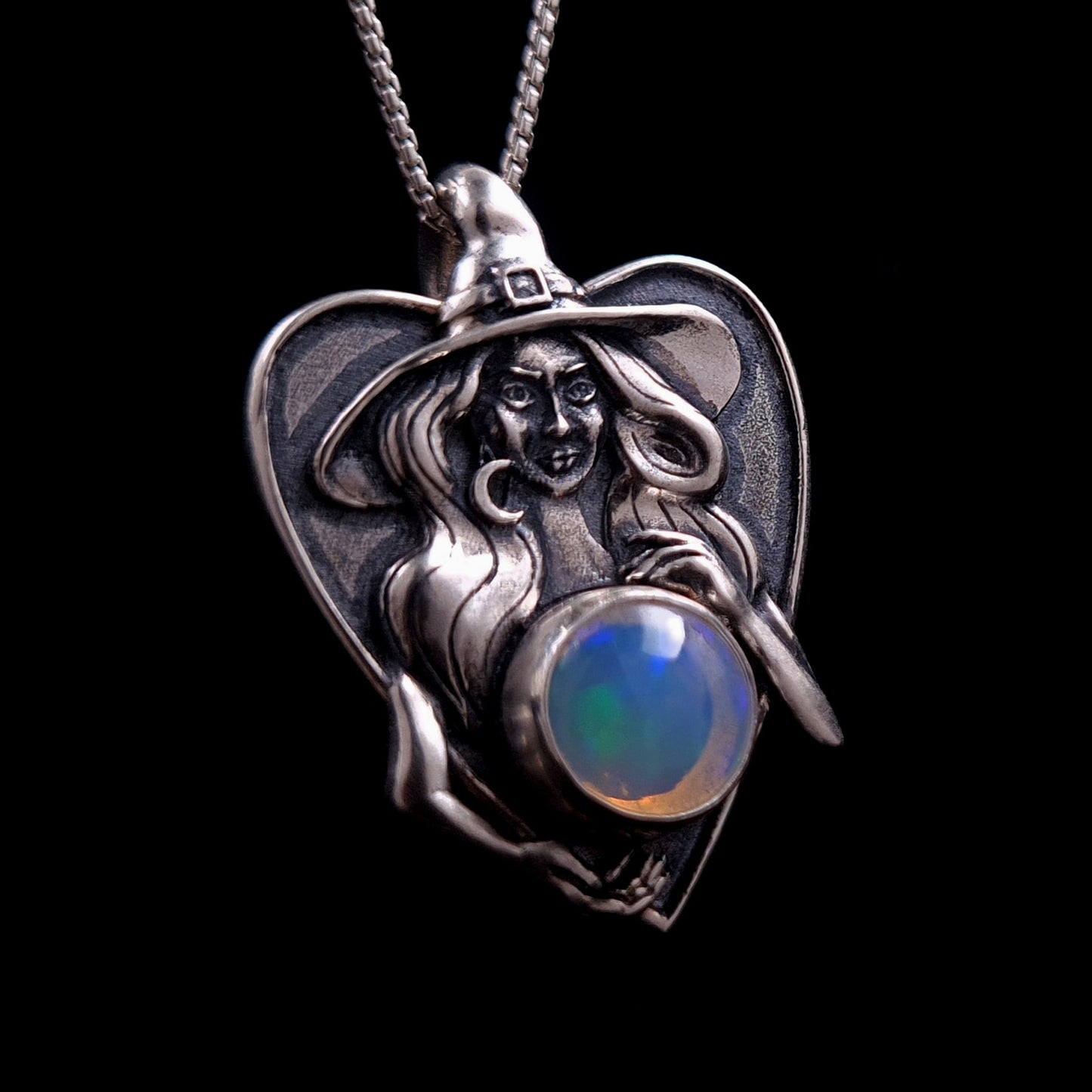 Frontal right-angled view of the sterling silver Crystal Ball Witch pendant by RBZ Jewelry. The sculptural Gothic witch figure is shown cradling a 10mm Ethiopian opal crystal ball with a glowing green and blue fire. Detailed features include the integrated bail in her pointed hat and a delicate crescent moon earring, set against a black background.