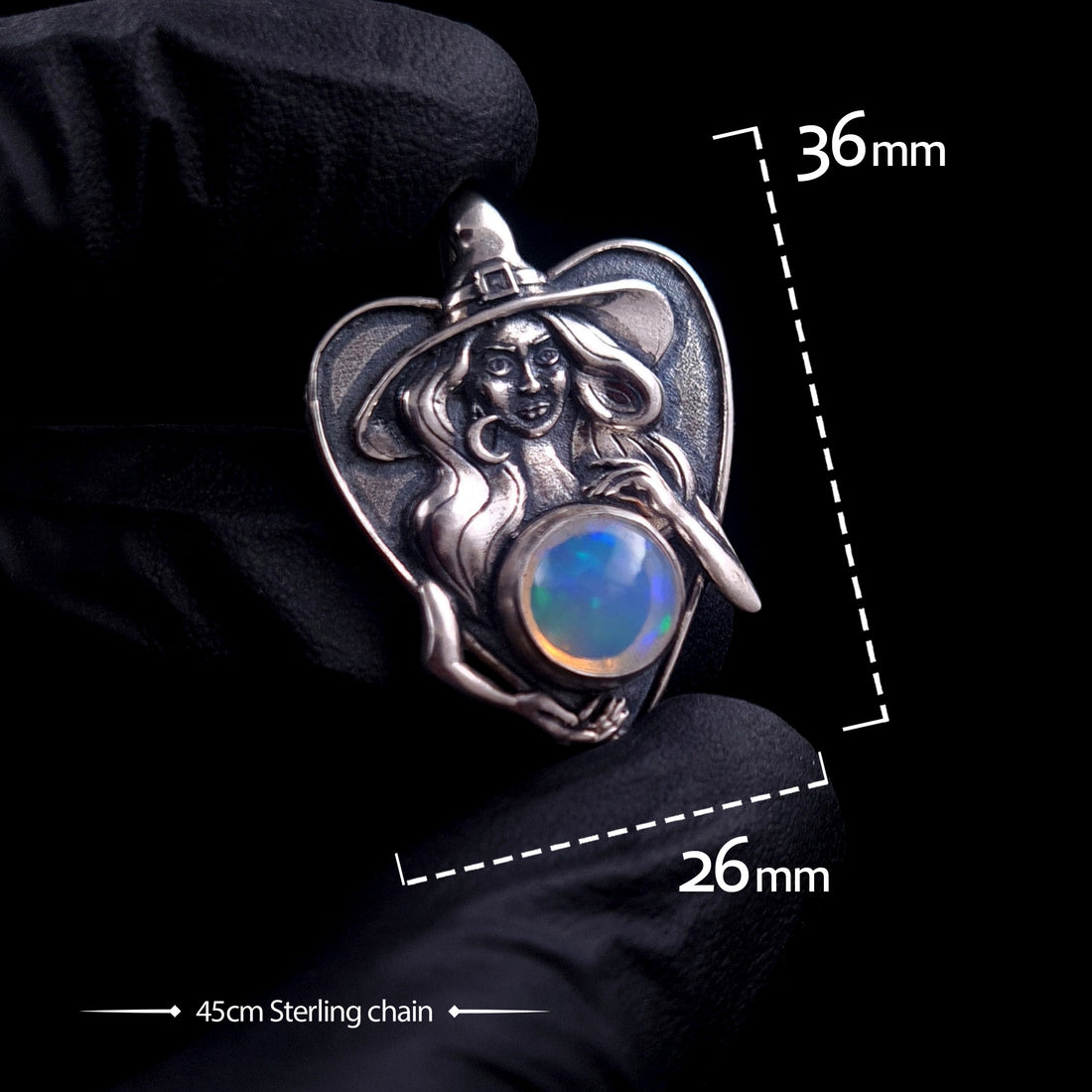A hand in a black glove holds the sterling silver Crystal Ball Witch pendant by RBZ Jewelry to show scale. White dimension overlays indicate a height of 36mm and a width of 26mm, with a caption for a 45cm sterling chain. The pendant features a detailed sculptural witch cradling a glowing 10mm Ethiopian opal against a black background.