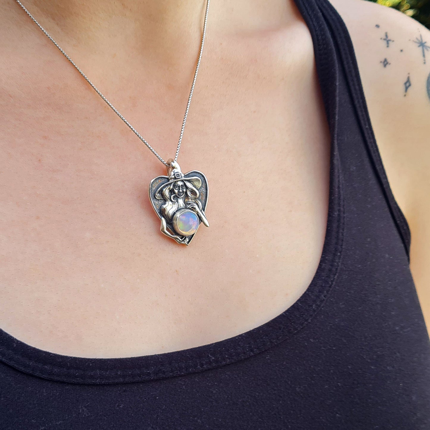 A lifestyle shot of a female model wearing the handcrafted sterling silver Crystal Ball Witch pendant by RBZ Jewelry. The pendant, featuring a sculptural witch figure cradling a 10mm Ethiopian opal, hangs from a fine silver chain against a black tank top, showcasing the jewelry's scale and how it sits when worn.