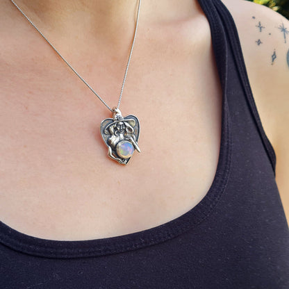 A lifestyle shot of a female model wearing the handcrafted sterling silver Crystal Ball Witch pendant by RBZ Jewelry. The pendant, featuring a sculptural witch figure cradling a 10mm Ethiopian opal, hangs from a fine silver chain against a black tank top, showcasing the jewelry's scale and how it sits when worn.