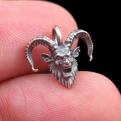 A close-up front view of the RBZ Jewelry Gothic Sterling Horned Demon pendant, held carefully between a person's thumb and index finger to demonstrate its intricate, small size and scale.