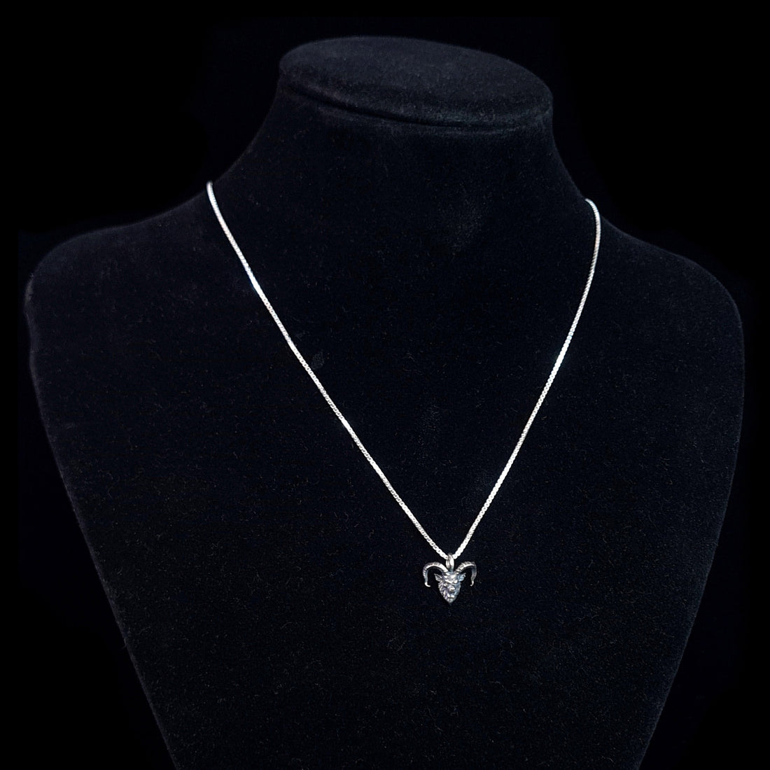 RBZ Jewelry Gothic Sterling Horned Demon pendant displayed on a black velvet neck mannequin to show how the piece hangs when worn. The small, oxidized sterling silver demon head with its signature snarling expression and curved horns is suspended from a bright silver chain against a solid black background.