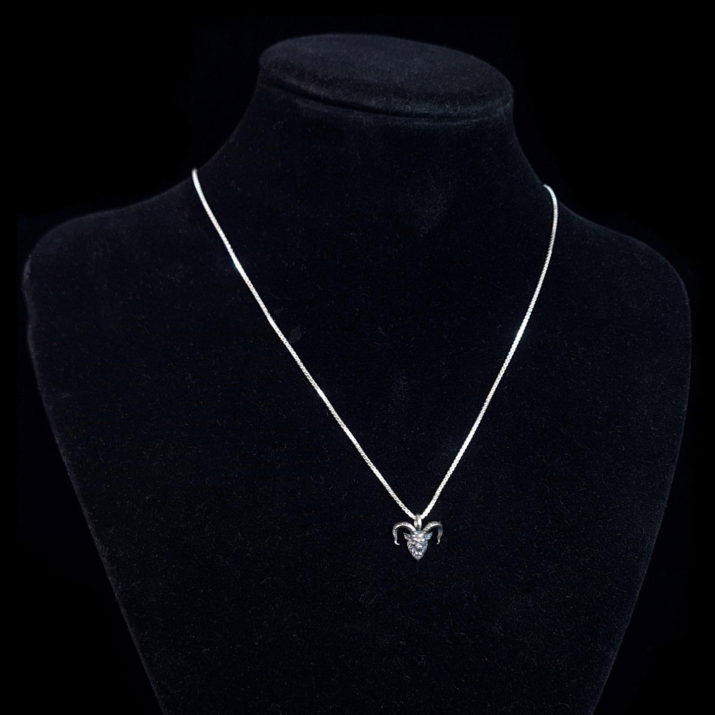 RBZ Jewelry Gothic Sterling Horned Demon pendant displayed on a black velvet neck mannequin to show how the piece hangs when worn. The small, oxidized sterling silver demon head with its signature snarling expression and curved horns is suspended from a bright silver chain against a solid black background.