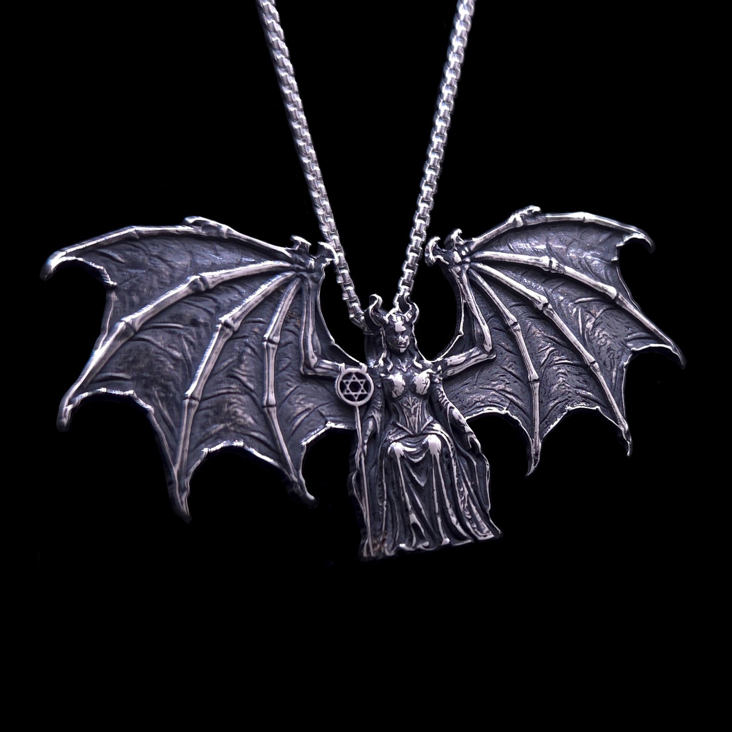 A highly detailed RBZ Jewelry Gothic Sterling silver pendant of the figure Lilith, who is enthroned between expansive, intricate, skeletal bat wings. The pendant features high-contrast dark oxidation, detailed armor on the figure, and a small scepter held in her right hand topped with a horned Star of David. Hanging from a silver chain against a solid black background.
