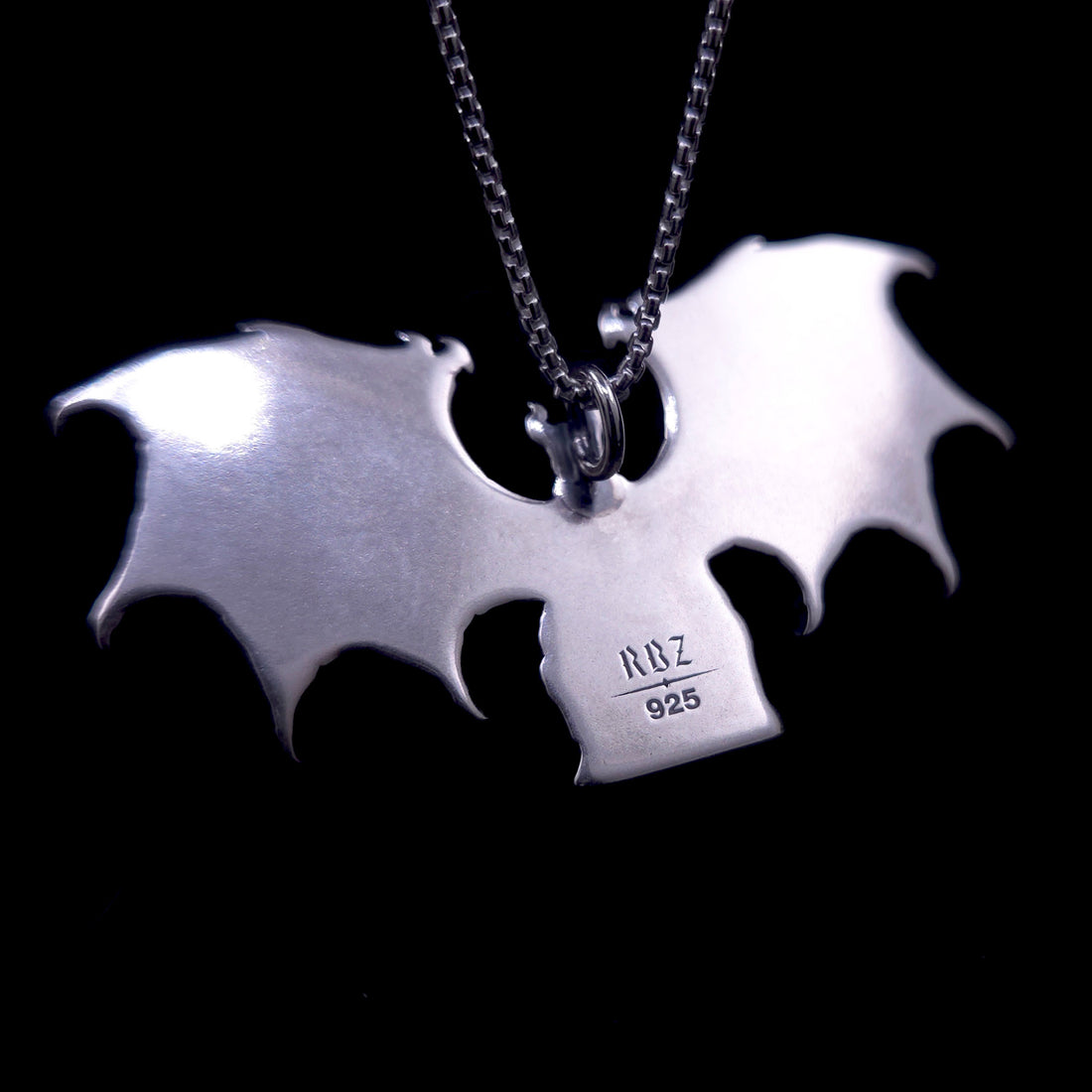 A detailed photograph of the reverse side of the RBZ Jewelry Gothic Sterling silver Lilith Demon Angel pendant, displaying a smooth, mirror-polished surface on the back of the enthroned figure and the expansive bat wings. Stamped clearly onto the back of the central body are the official RBZ hallmark and the '925' sterling silver purity mark. A sterling silver chain is attached at the top loop, with the pendant set against a deep black background.