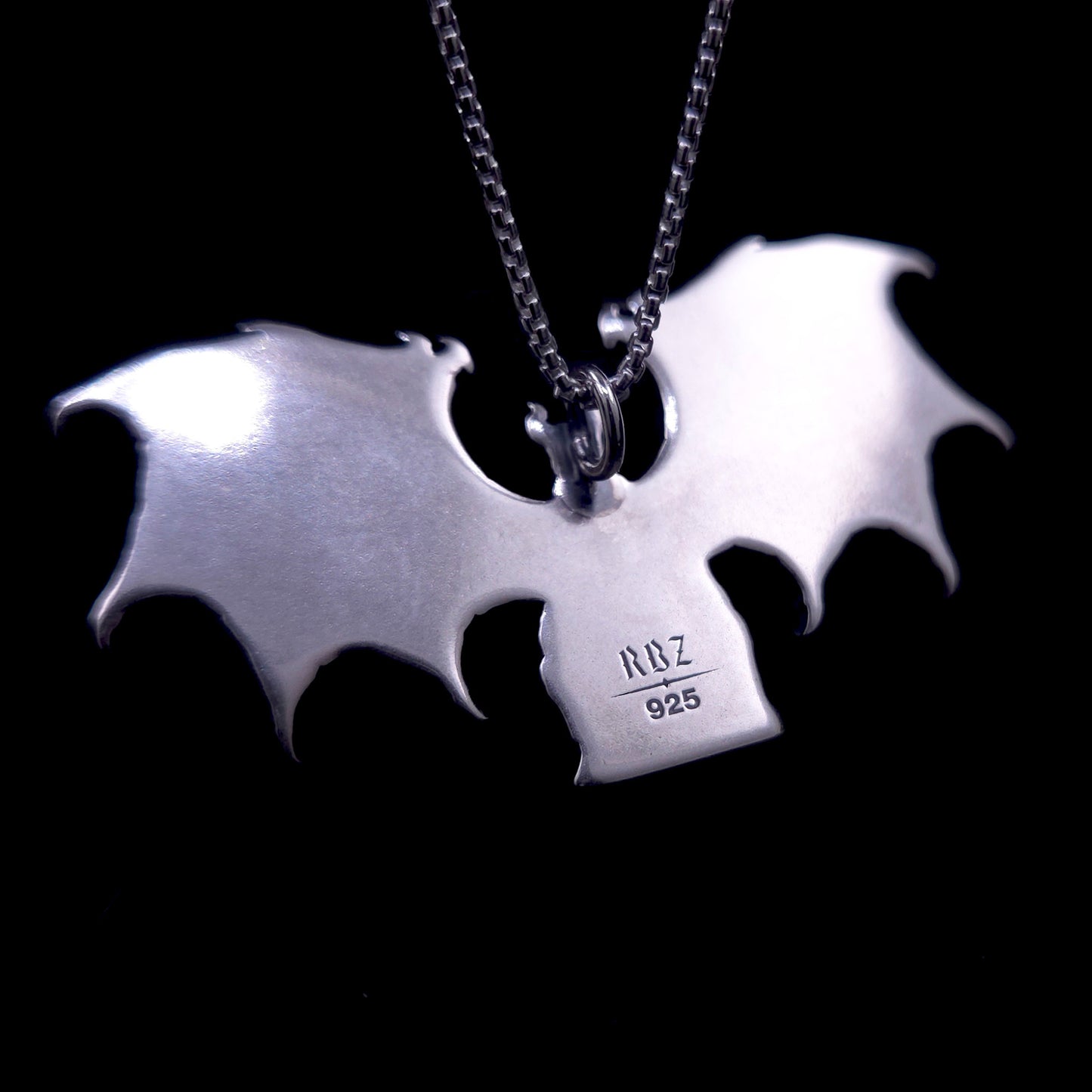 A detailed photograph of the reverse side of the RBZ Jewelry Gothic Sterling silver Lilith Demon Angel pendant, displaying a smooth, mirror-polished surface on the back of the enthroned figure and the expansive bat wings. Stamped clearly onto the back of the central body are the official RBZ hallmark and the '925' sterling silver purity mark. A sterling silver chain is attached at the top loop, with the pendant set against a deep black background.