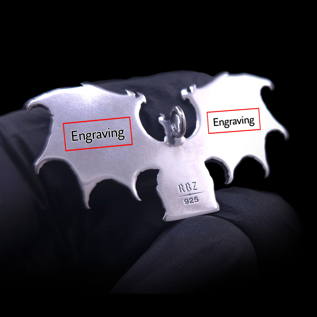 A detailed close-up shot of the polished reverse of the RBZ Jewelry Gothic Sterling silver Lilith Demon Angel pendant, held in a black-gloved hand. Red-outlined rectangular boxes are superimposed over the back of the left and right skeletal bat wings, each labeled with the word 'Engraving' in black text, illustrating the customizable areas. The existing 'RBZ' hallmark and '925' stamp are visible on the figure's base against a dark background, allowing for personalized text and messaging.