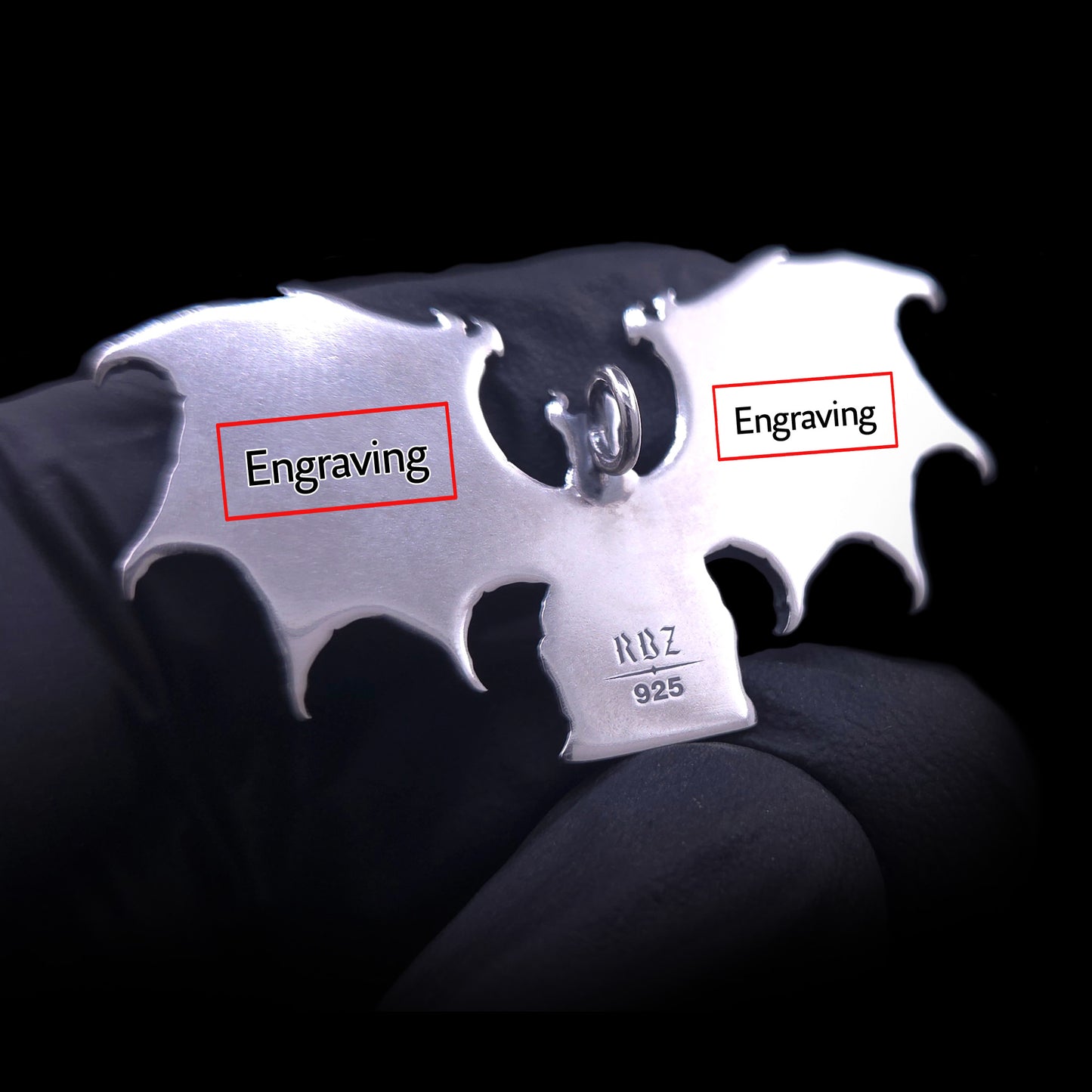 A detailed close-up shot of the polished reverse of the RBZ Jewelry Gothic Sterling silver Lilith Demon Angel pendant, held in a black-gloved hand. Red-outlined rectangular boxes are superimposed over the back of the left and right skeletal bat wings, each labeled with the word 'Engraving' in black text, illustrating the customizable areas. The existing 'RBZ' hallmark and '925' stamp are visible on the figure's base against a dark background, allowing for personalized text and messaging.