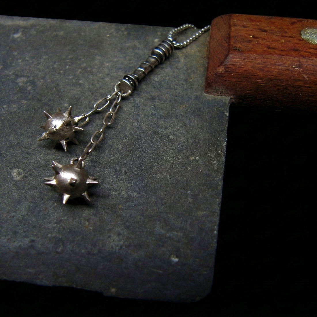 Sterling silver Flail Crusher pendant by RBZ Jewelry, featuring two detailed spiked mace spheres connected by chains, resting on the worn metal blade of a butcher's knife, embodying a raw, medieval war relic aesthetic.
