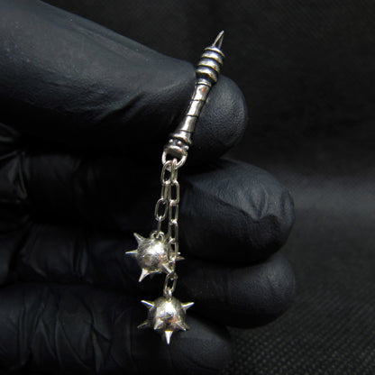 Sterling silver Flail Crusher pendant by RBZ Jewelry, held in a black gloved hand against a dark background, displaying the miniature artifact-like handle with spiral grip and the duo of hand-soldered spiked mace spheres on chains.