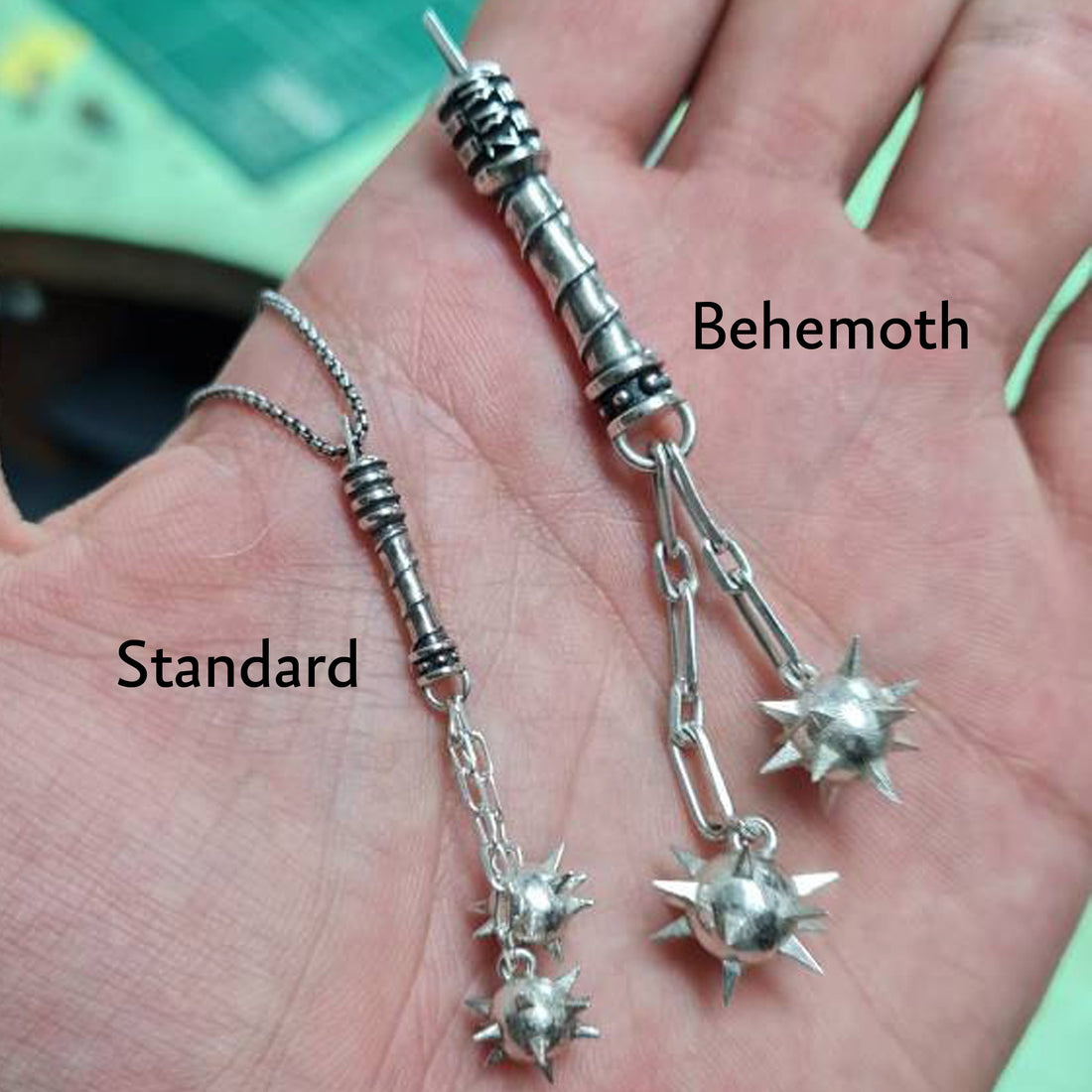 Size comparison of the Sterling silver Flail Crusher pendant by RBZ Jewelry, showing the Standard (45mm) and Behemoth (76mm) versions held in a hand to demonstrate scale, both featuring handcrafted spiked mace spheres and spiral-textured handles.