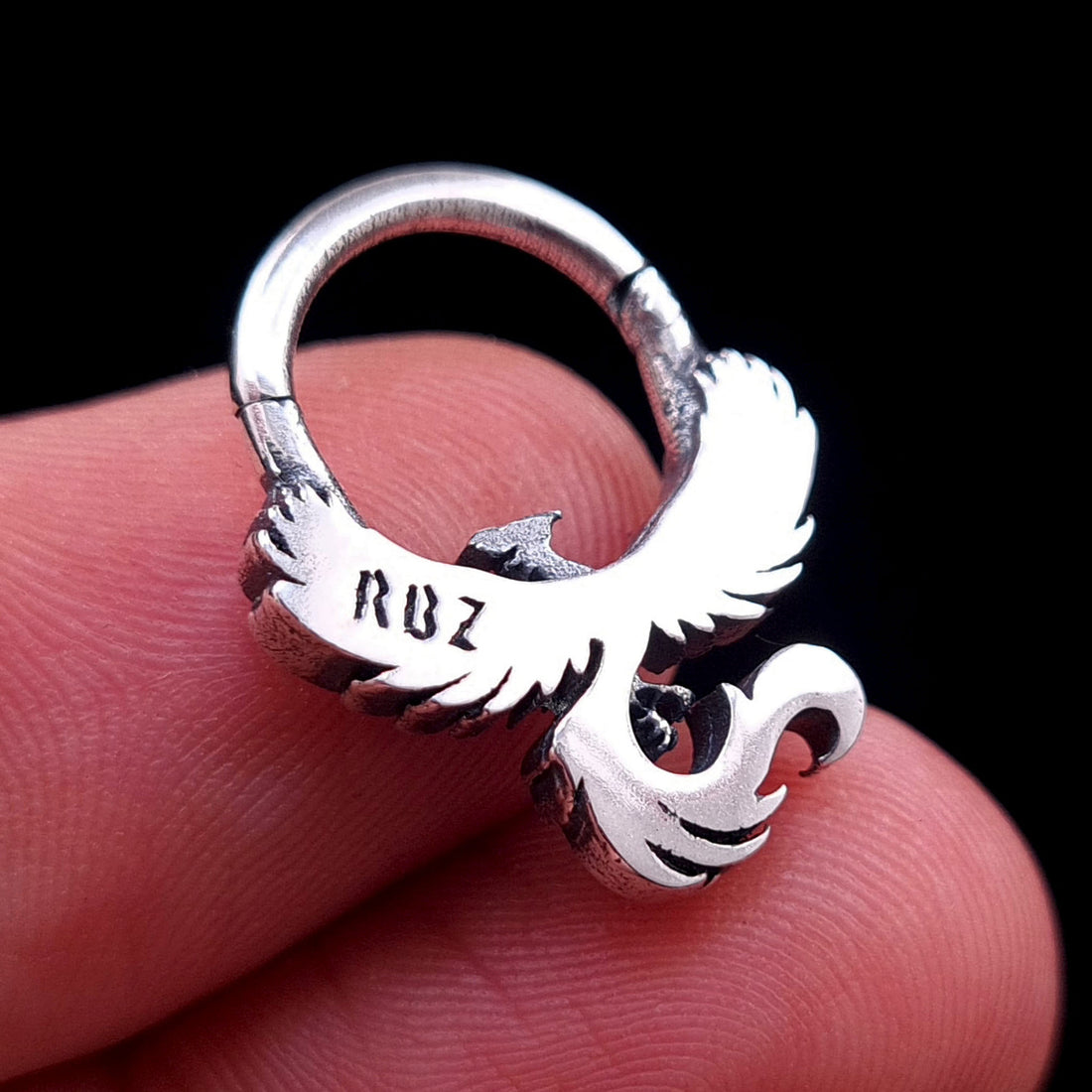 Close-up back view of the sterling silver Soaring Phoenix septum ring from RBZ Jewelry, held between fingertips to show scale and the smooth, polished back surface with the prominently stamped 'RBZ' maker's mark against a black background.