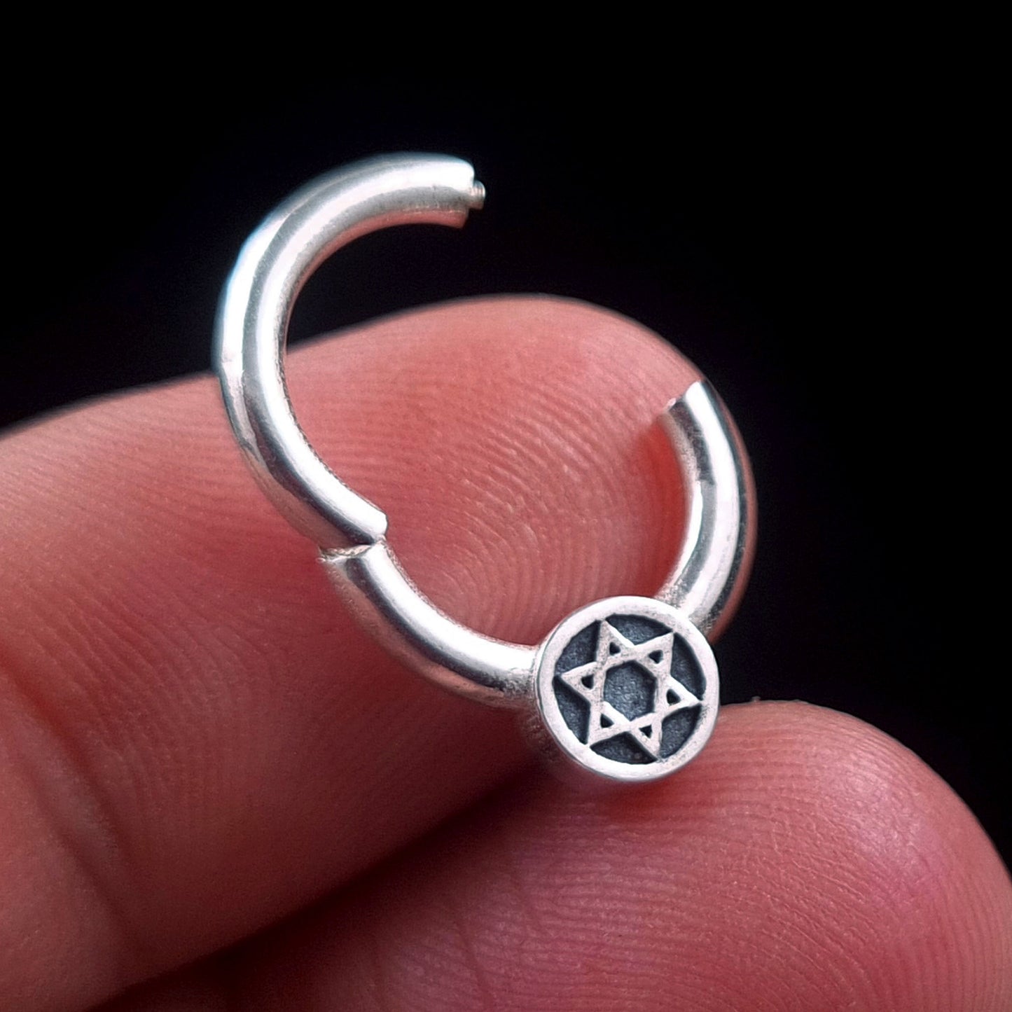 A close-up photograph of the Sterling silver Star of David septum clicker ring by RBZ Jewelry, held in an open position between a human thumb and index finger against a solid black background. The hinged section of the ring is unlatched, demonstrating its function, with the Magen David medallion clearly visible on the lower part of the ring.