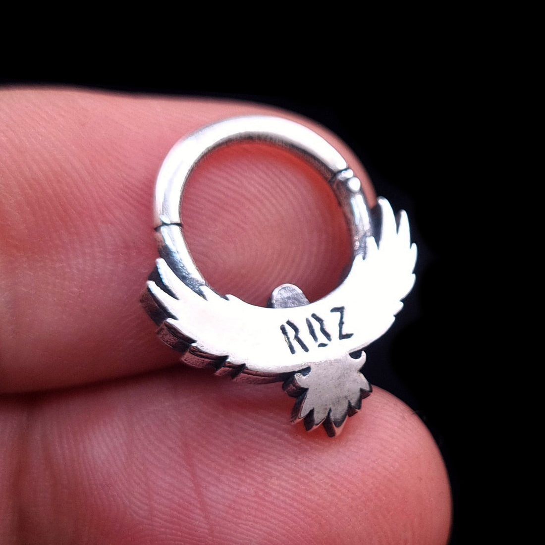 Close-up back view of the Sterling silver Tribal Eagle septum ring by RBZ Jewelry, held in hand to clearly display the engraved "RBZ" logo hallmark on the eagle silhouette and the clicker hinge mechanism, against a black background.