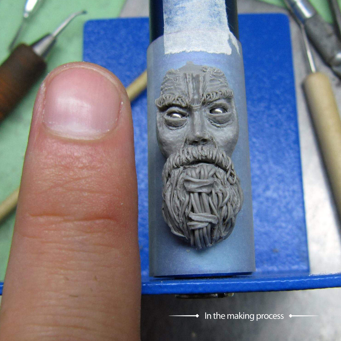 A close-up of the hand-sculpted master model for the RBZ Jewelry Viking warrior ring during the jewelry making process, showing the intricate facial and beard details in gray sculpting material next to a thumb for scale.