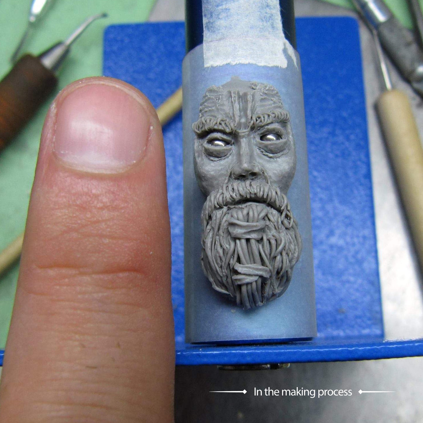 A close-up of the hand-sculpted master model for the RBZ Jewelry Viking warrior ring during the jewelry making process, showing the intricate facial and beard details in gray sculpting material next to a thumb for scale.