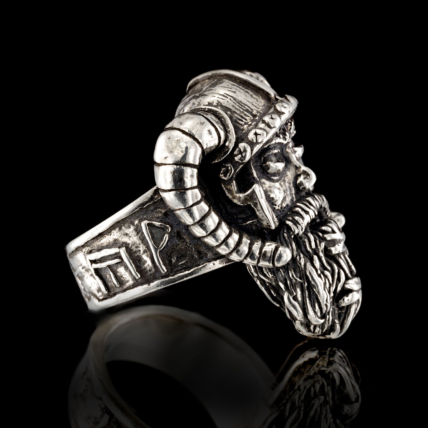 A macro view of the left-hand side profile of the RBZ Jewelry Sterling silver Viking Warrior ring. This detailed perspective focuses on the intricate outer shank band, which prominently displays a sequence of four hand-sculpted runic inscriptions, including the Axe-Hammer (Mjöllnir), and specific symbols for Joy, Wealth, and Giving, as described in the product details. The detailed left-side profile of the weathered warrior’s face and beard is also clearly visible. The aged silver ring rests on a black BG.